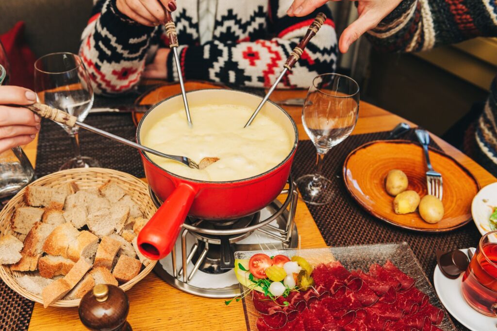 A red fondue pot filled with melted cheese and three fondue forks resting inside, along with a table laid with bread and cold cuts.