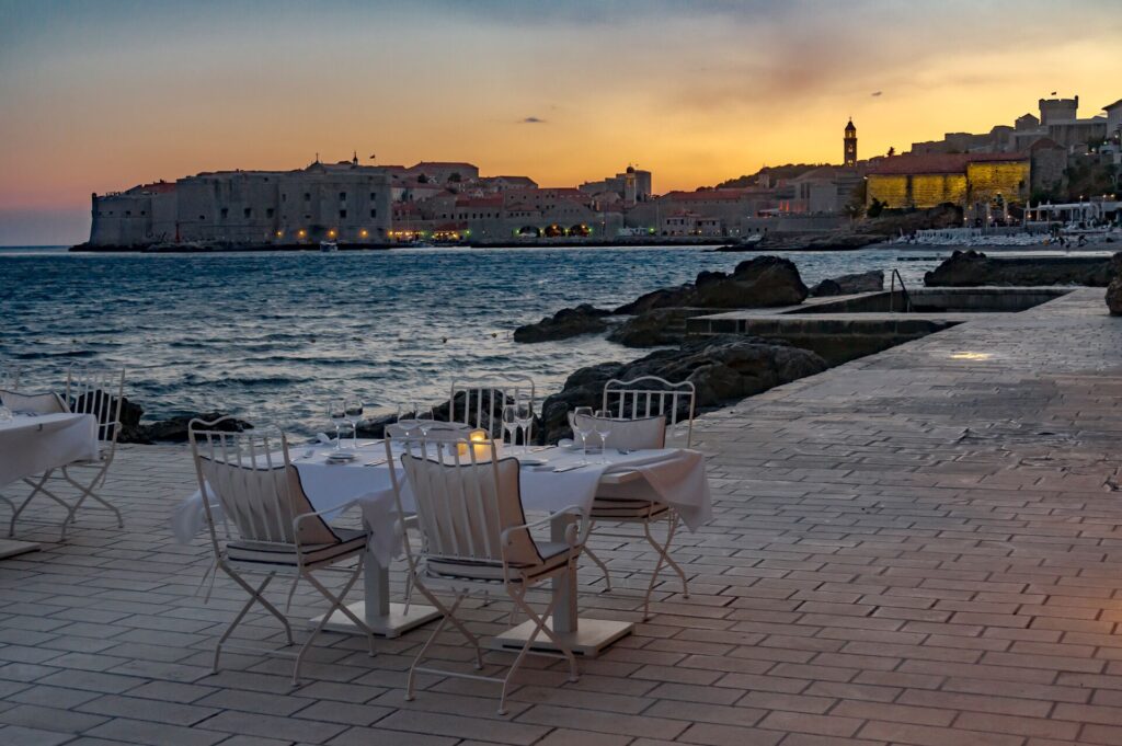 A table and chairs set for dinner on a promenade by the sea in Dubrovnik, with a sunset in the background