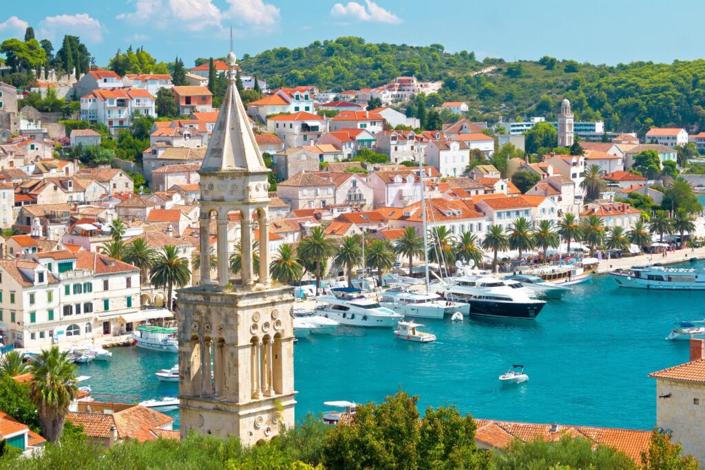 Red roofs and white buildings line the harbour in Hvar with bright blue water and a chruch townr in the foreground. 