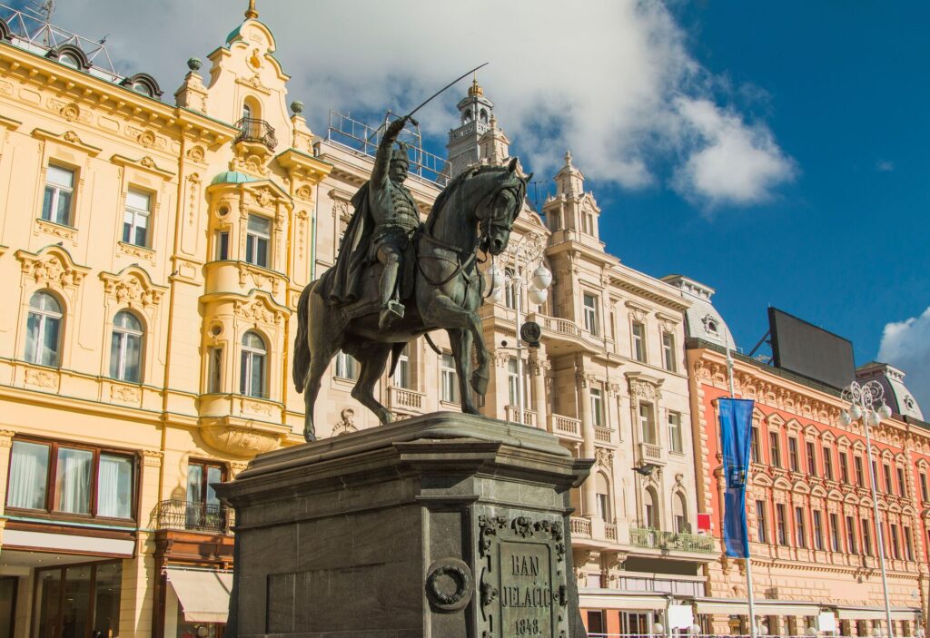 A soldier sits atop a horse as the Ban Jelacic statue on Jelacic Square in Zagreb, with grand buildings in the background in pale yellow, pink and peach.