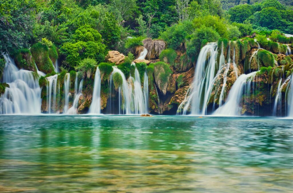 Cascading waterfalls into a large green lake wit bright green vegetation in the background at Kyka National Park in Croatia.