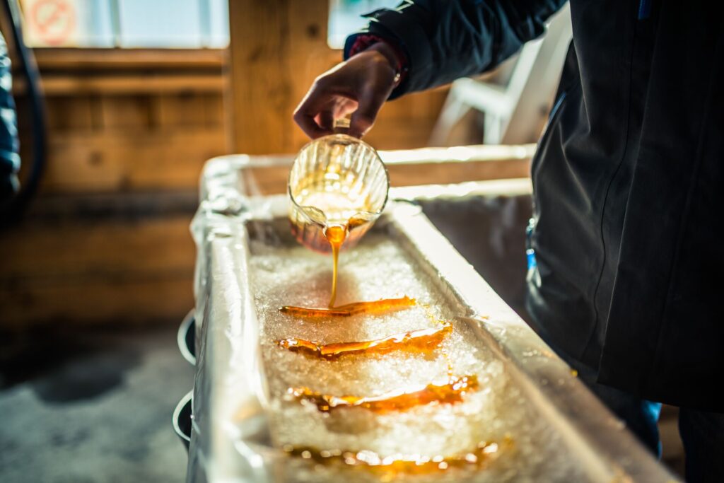 Bright orange maple syrup is poured by a hand from a glass jug onto white snow, with a blurred background