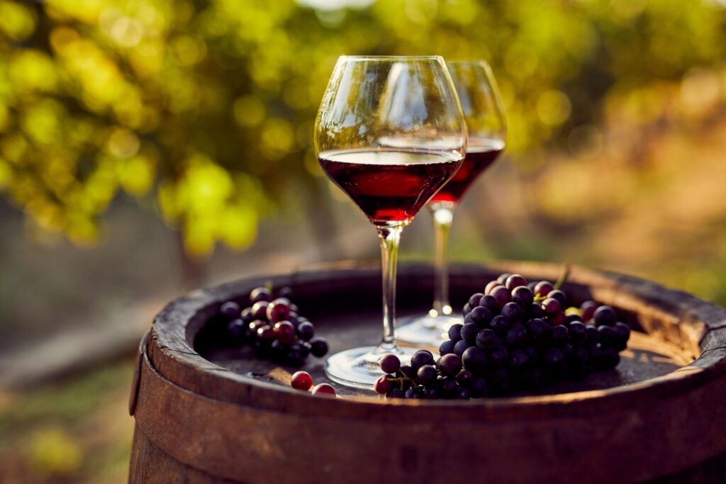 Two glasses of red wine sit atop a dark brown wood barrel, with red grapes and a blurred background of green vines.