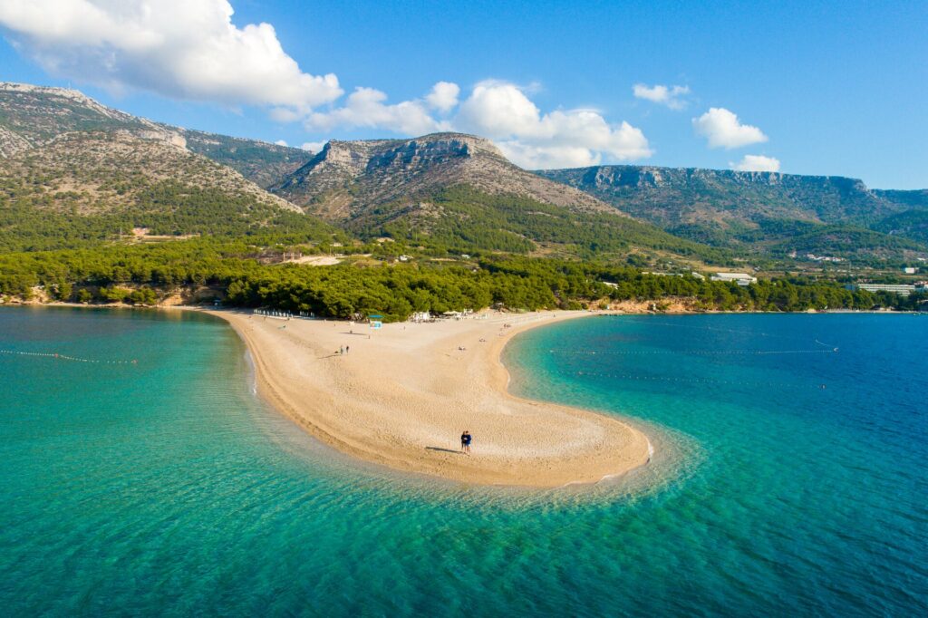 A golden beach juts out into bright azure waters with green forrest and mountains behind.