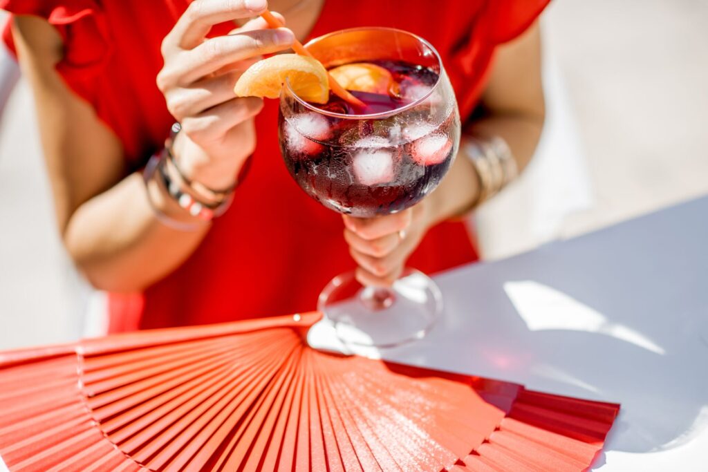 Close up of a woman holding a glass of a bright red drink with ice cubes, wearing a red dress with a red Spanish fan on the table. 