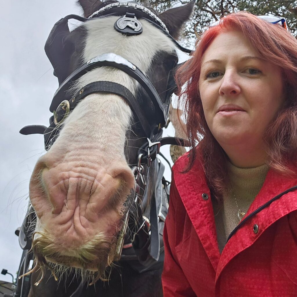 The head of a black and white horse wearing a bridle with silver badges on it is pictures next to Travel Director Erica who is wearing a bright red coat.