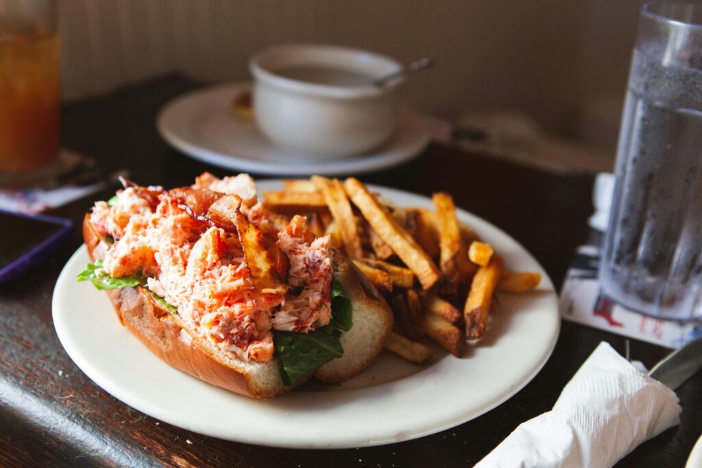 A white plate with a hot dog roll filled with lobster and a side of chips is shown on a brown table with a glass of water beside