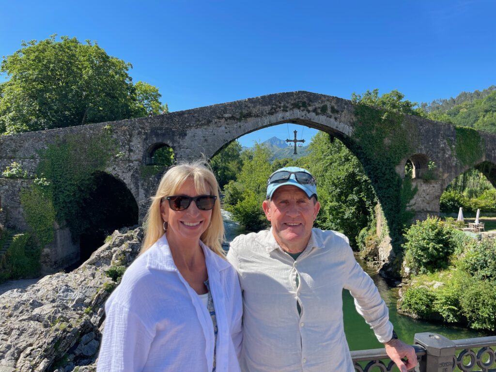 A man and a woman stand side by side smiling, with an ancent grey stone bridge behind, with green vegetation and a bright blue sky