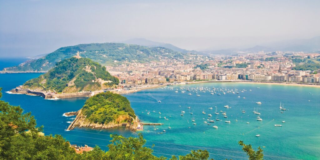 Aerial shot of bright blue waters, green islands and the town of San Sebastian in Spain with a long sandy beach, all bathed in sunlight