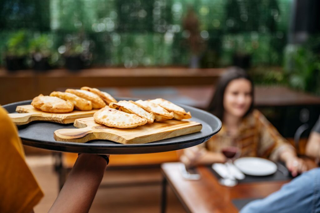 A plate of golden emapanadas is carried to an outdoor wooden table where a lady is seated