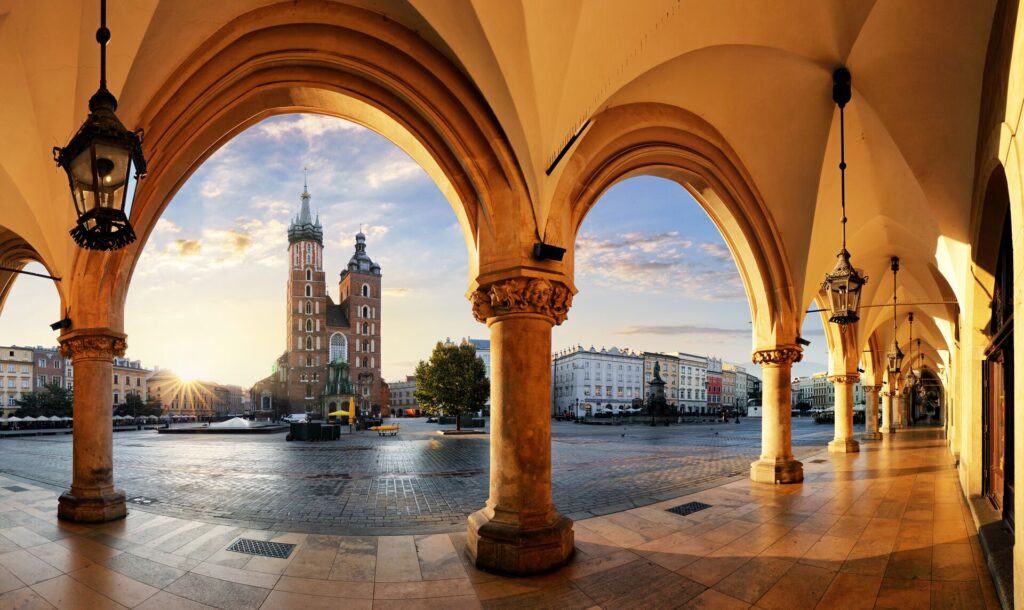 A tall tower building in Krakow is pictured through semi circle struts of another building, with the sun shining behind.