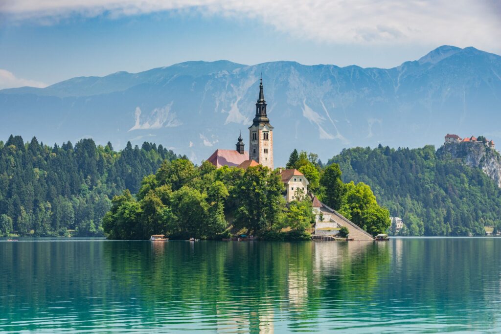 A tower and buildings on a small island with bright green trees, on a blue lake with mountains behind in Slovenia.