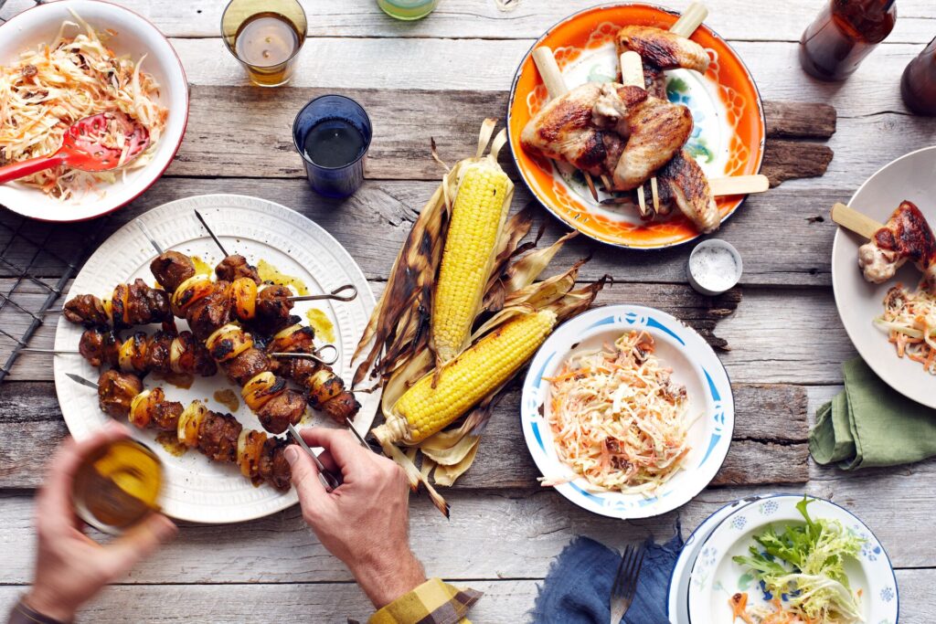 Aerial shot of a table with colorful plates of BBQ food, including corn on the cob, meat kebabs and coleslaw.