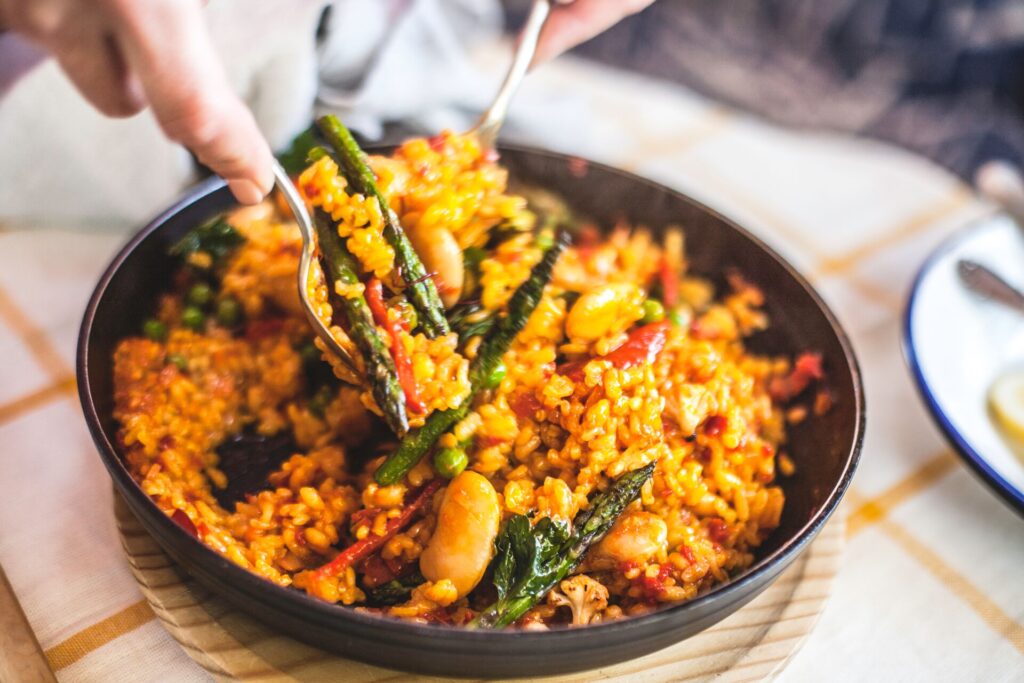 A close up of Paella with rice and red and green vegetables being spooned in an iron pan