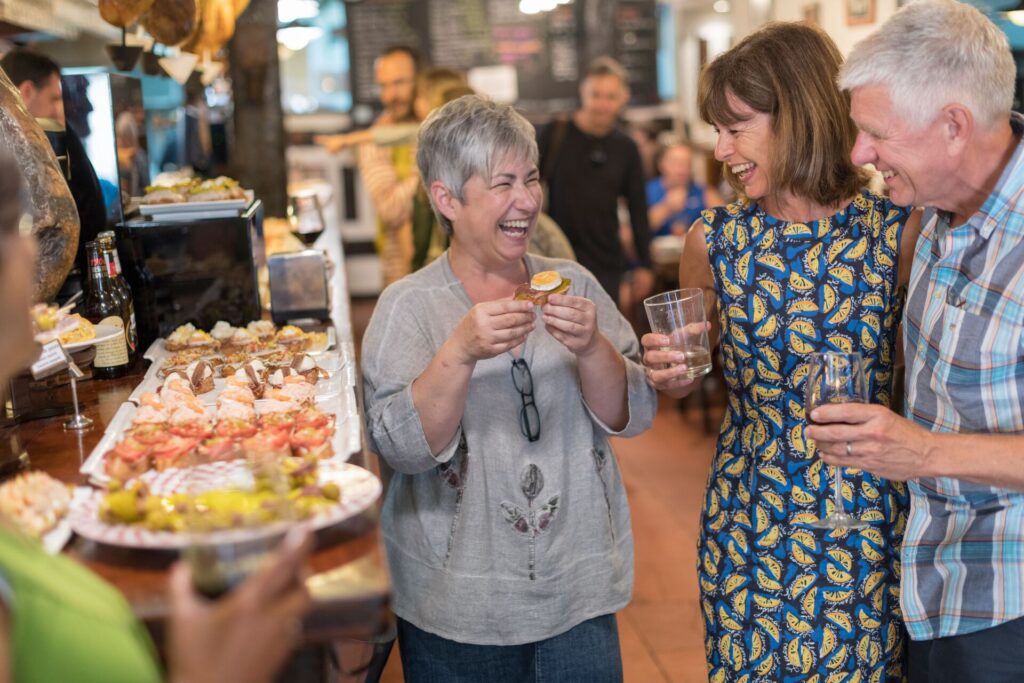 Guests laugh and smile in a Portuguese food market, with plates of pinxos, small pieces of finger food, beside them.