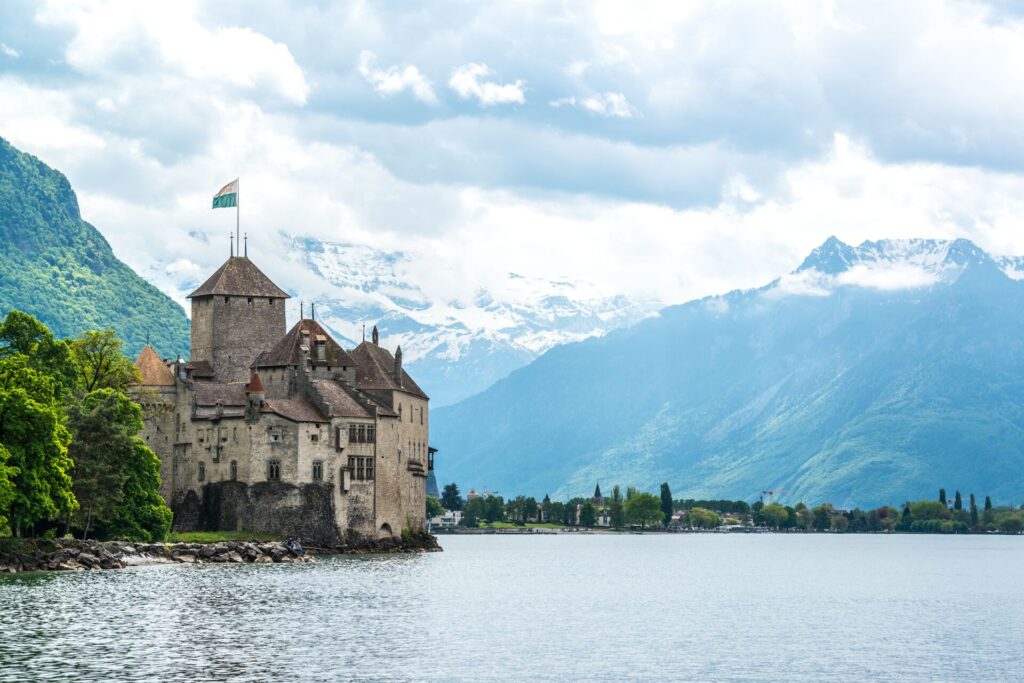 A brown and beige brick castle perched on a rock on the shore of a large lake with snow capped mountains in the background.