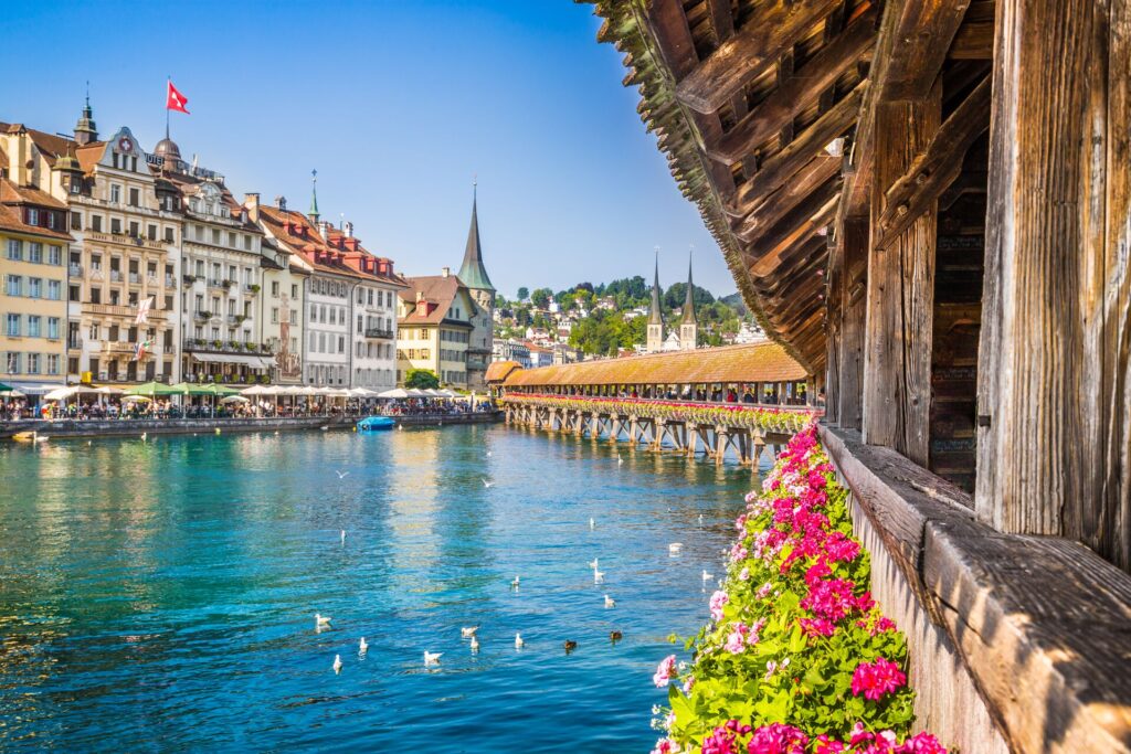 Lucern's ancient bridge with pink flowers and blue waters, with buildings in the background.