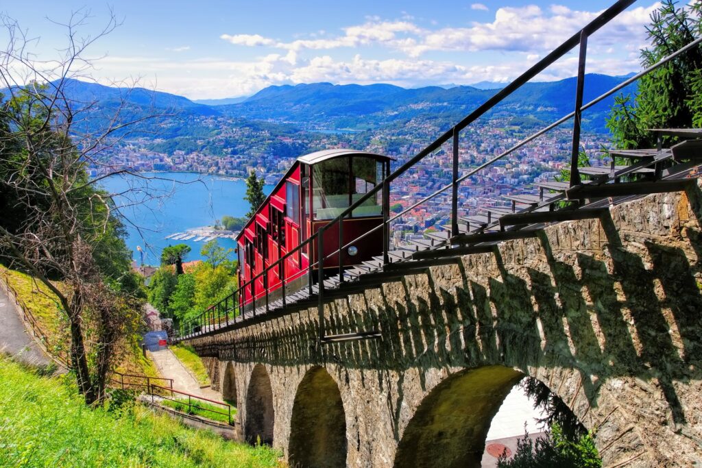 A bright red funicular climbs a stone viaduct type track, with a blue lake, mountains and a sprawling city in the background.