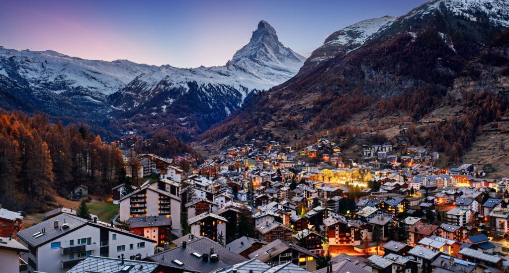 The alpine town of Zermatt in the evening with lights on in houses, flanked by snow covered Alps and the pointed Matterhorn mountain. 