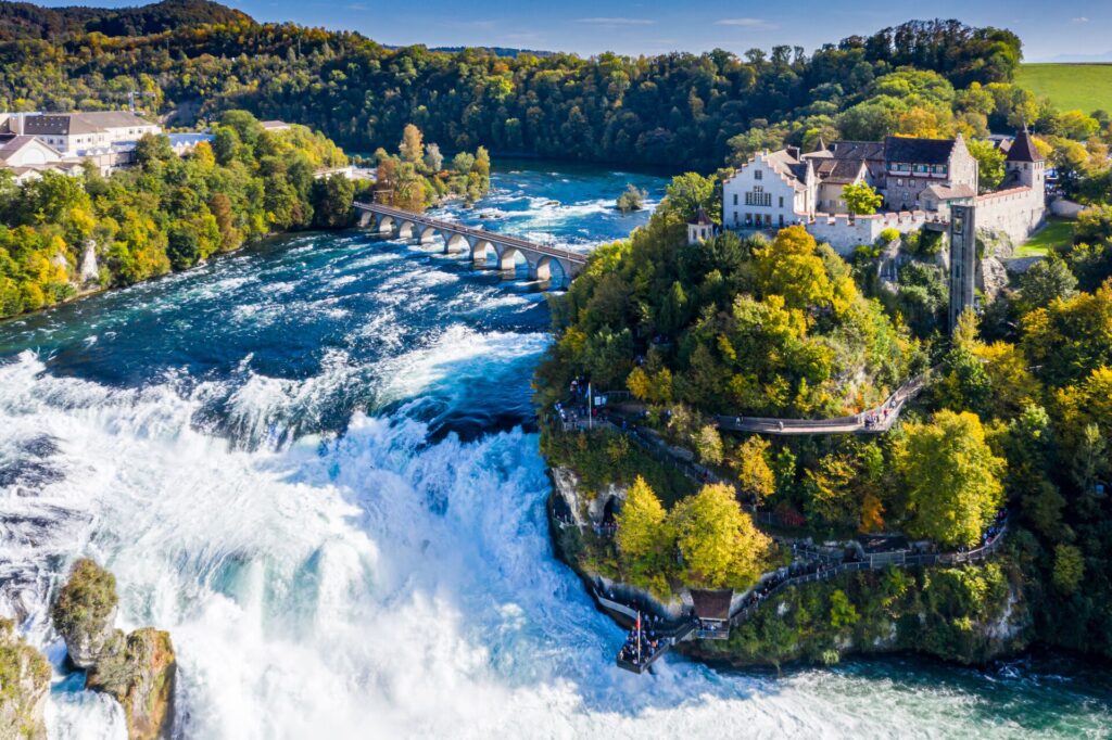 Flowing waters and cascades of the Rhine Falls in Switzerland, with green trees on the banks and ancient buildings.