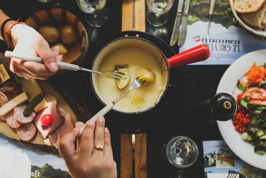 Aerial shot of a traditional Swiss fondue pot filled with melted Gruyere cheese, and peoples hands as the dip in potatoes.