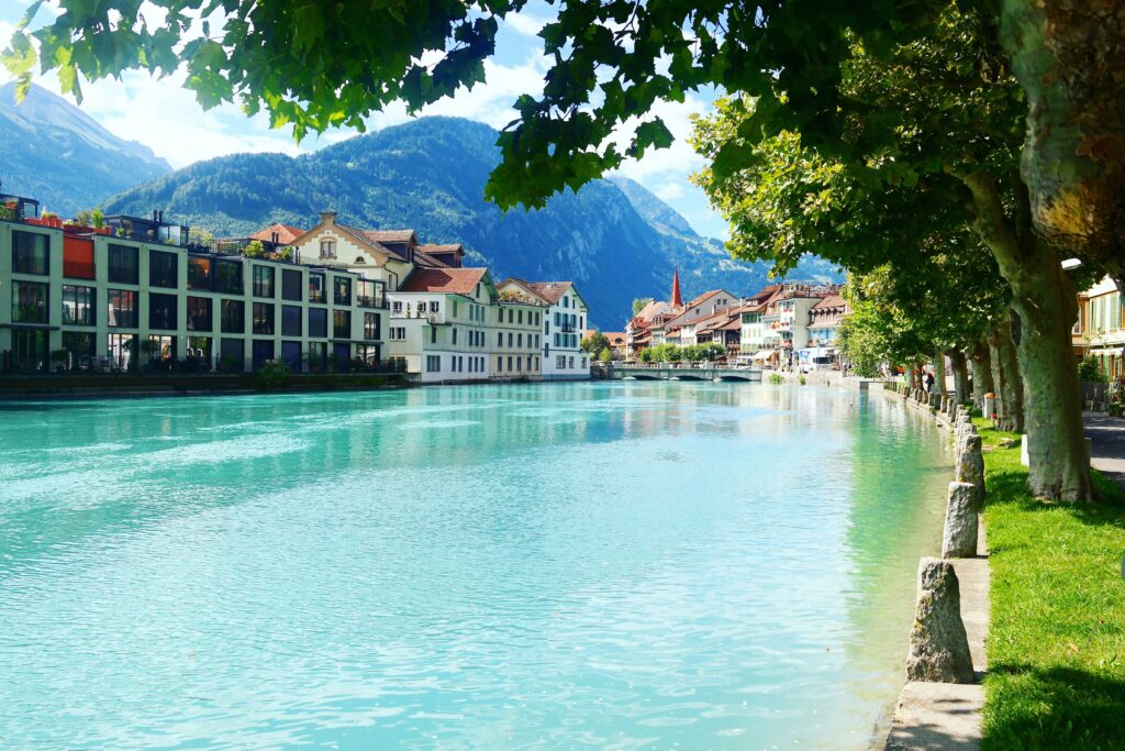 Buildings sit alongside a bright blue lake water with trees alongside and mountains in the background.