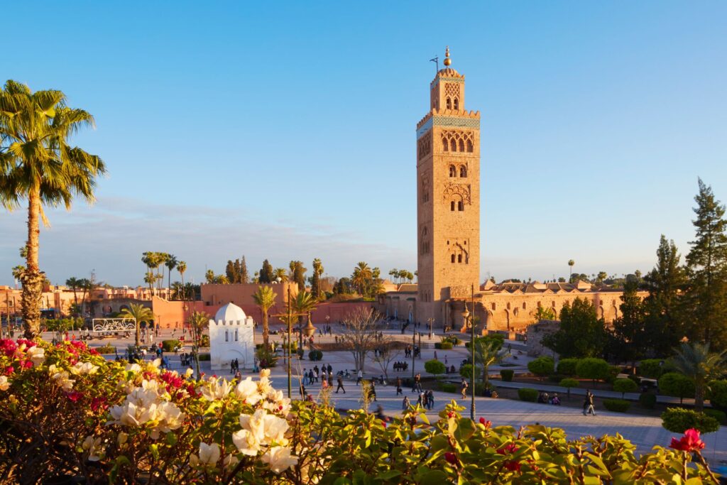 A tall mosque tower is surrounded by buildings, open square and flowers and trees.