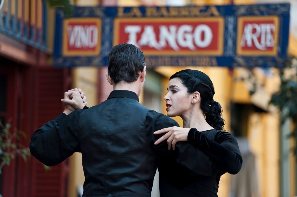 A man and woman dance Tange in front of a red sign saying Tango.