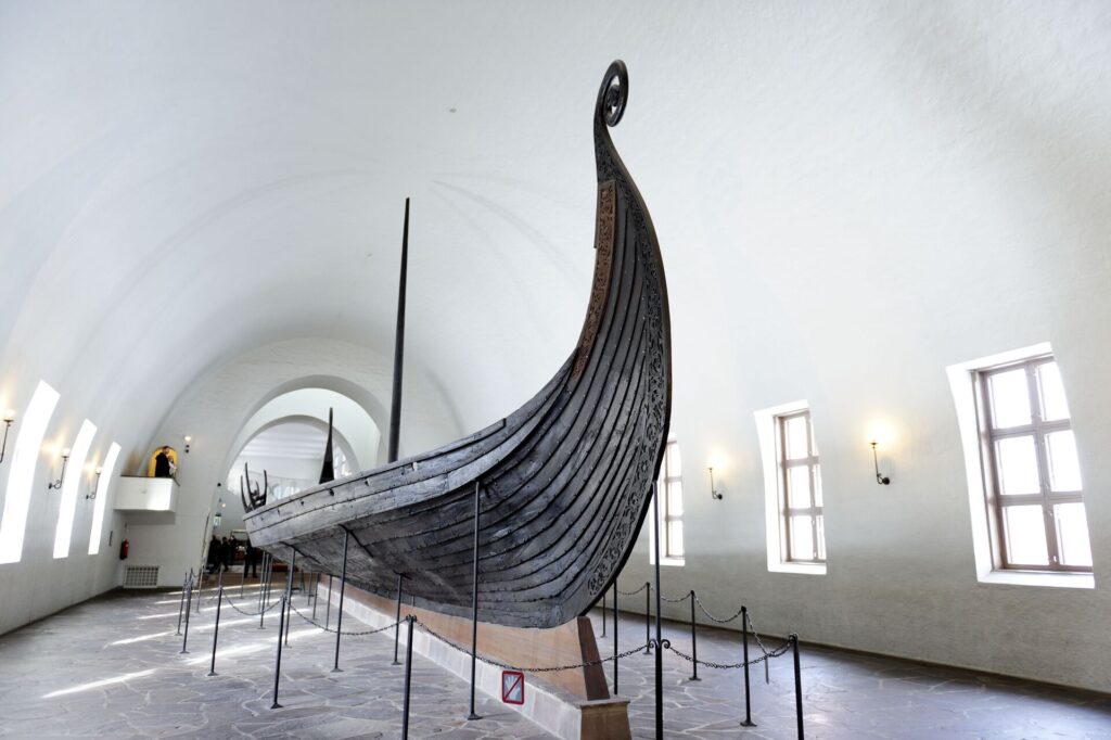 An ancient dark brown wooden Viking boat in a museum with white walls and windows in Oslo, Norway.