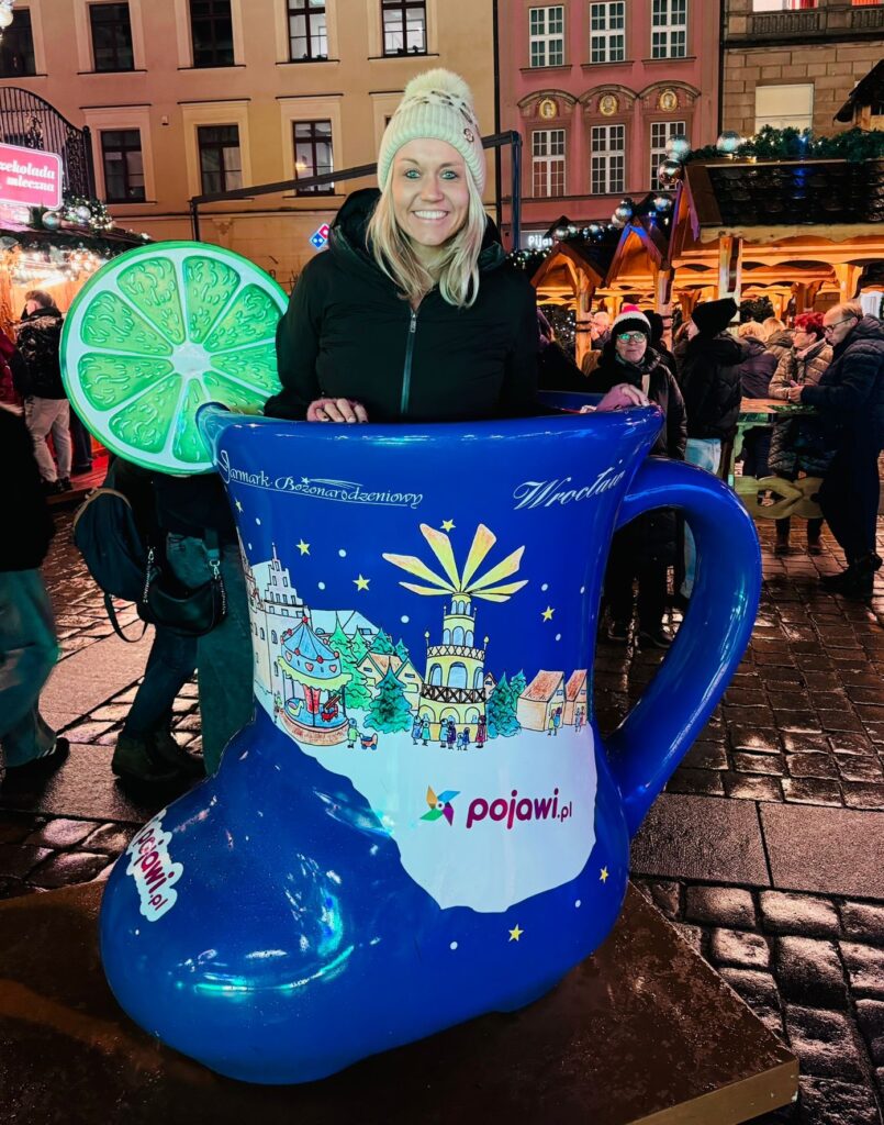 Travel Expert Dani stands in a large blue boot in a Christmas Market in Europe, with blonde hair, a hat and a smile