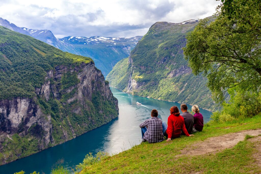 Four friends over look Geirangerfjord in Norway, with deep blue water and mountainous sides.