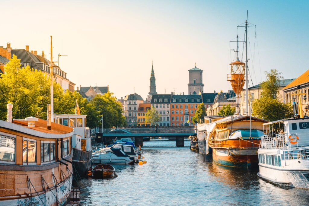 Moored boats on either side of a blue water canal, with orange and shite coloured houses with black roofs in Copenhagen, Denmark.