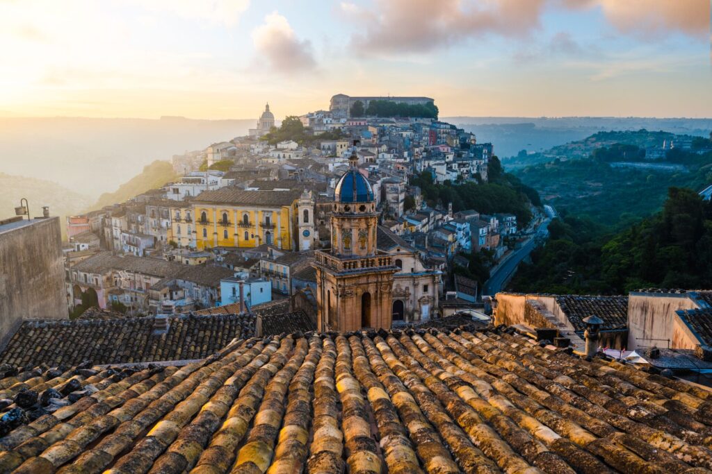 An old town in Sicily is shown from the view of looking down a sloping tiled roof, with the neon set on a hill, in low light.