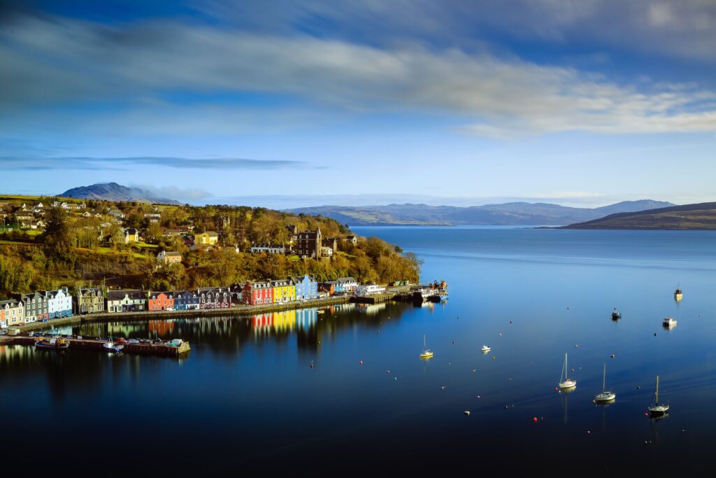 Dark bleach sea, with a row of yellow, blue, red and coloured houses on the seafront with green trees behind and mountains in the distance.