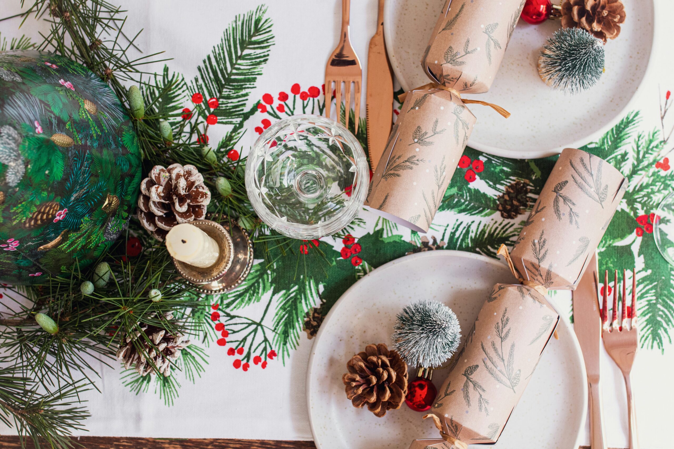 Aerial shot of a table decorated for Christmas with green holly, brown fir cones, a decorative napkin and plates and Christmas crackers.