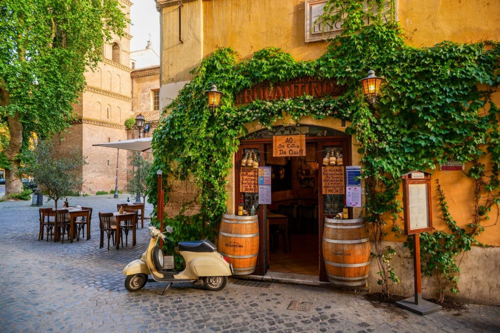 The front of small trattoria restaurant in Italy with wooden barrels outside, a menu and a scooter parked in front.