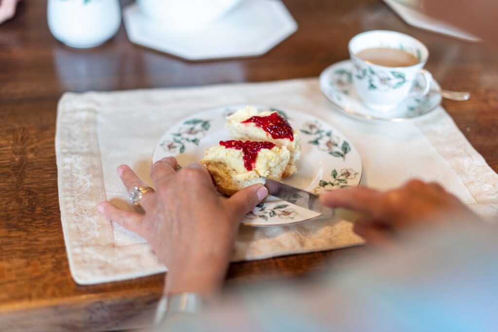 women's hands use a knife to cut into a scone laden with clotted cream and jam, on a decorative china plate with a cup of tea and saucer alongside.