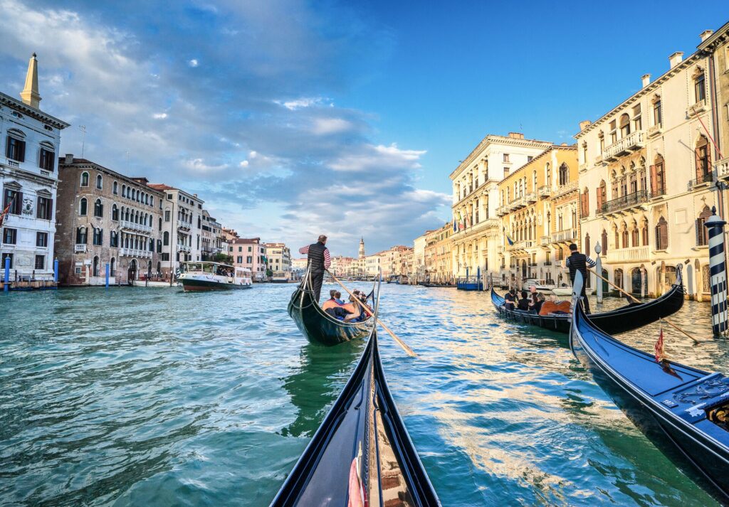 Gondolas with people inside float on the rover in Venice, Italy, flanked by old buildings and a blue cloudy sky.