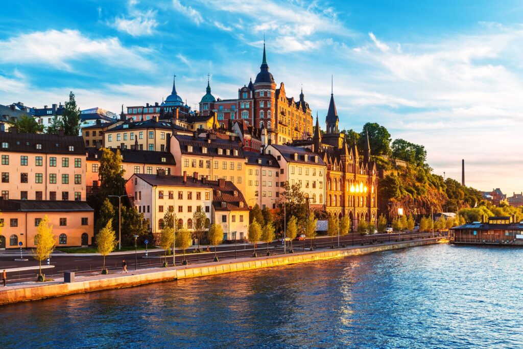 Buildings and a red coloured castle in Stockholm, Sweden shine in the evening lights, with autumn trees and a river alongside.