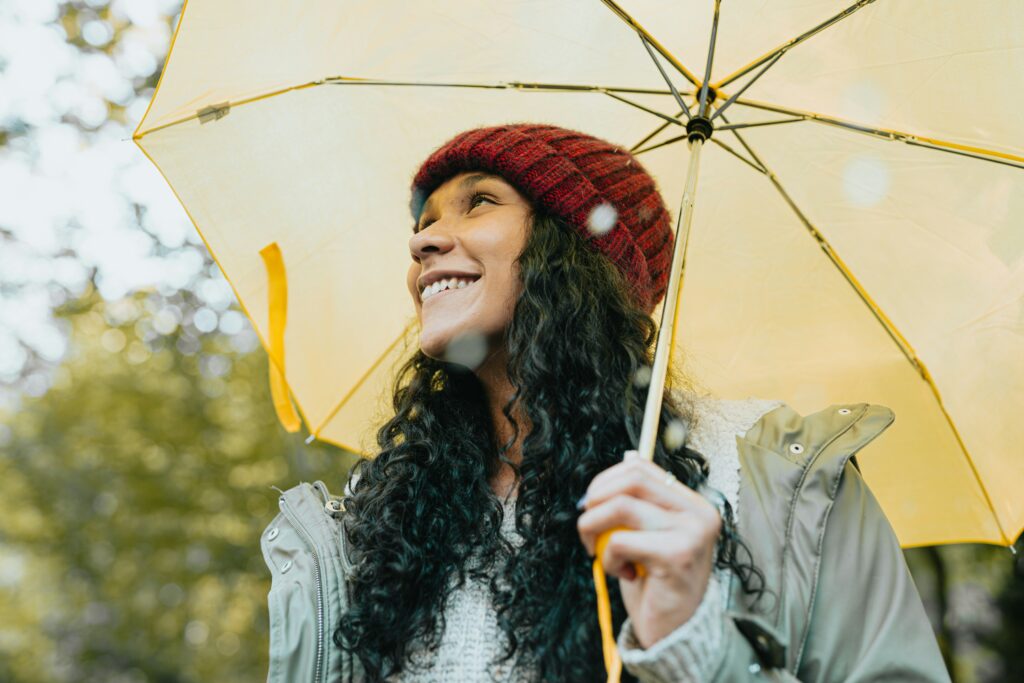 A girl with long black hair and a red hat carries a yellow umbrella with blurred trees and sky in the background.